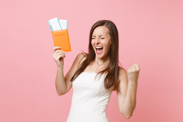 Joyful bride woman in white wedding dress doing winner gesture, holding passport and boarding pass ticket, going abroad for honeymoon, vacation isolated on pastel pink background. Wedding celebration.