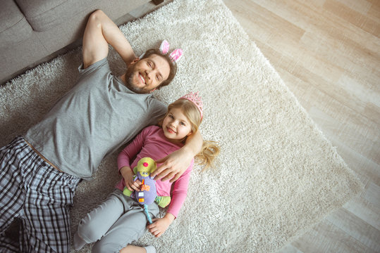 Relaxing at home together. Top view of joyful dad is lying of soft carpet and embracing his little daughter. They are looking at camera and laughing. Copy space