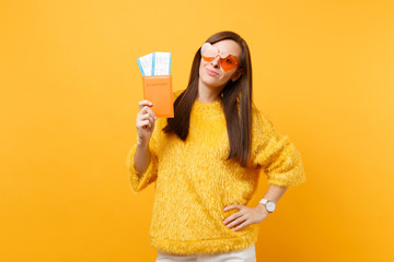 Attractive young woman in fur sweater, orange heart eyeglasses holding passport and boarding pass tickets isolated on bright yellow background. People sincere emotions, lifestyle. Advertising area.