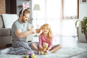 Caring father is combing hair of his little daughter. Cheerful girl is sitting on floor and smiling. She is holding teddy bear 