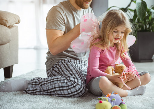 Pretty blond girl is playing with toys while her daddy is combing her hair. She is looking at pink crown dreamingly and smiling