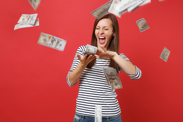 Crazy woman in striped clothes screaming scattering throwing out money banknotes lots of dollars isolated on bright red wall background. People sincere emotions, lifestyle concept. Mock up copy space.