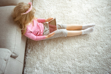 Top view of cheerful little girl is watching interesting video on tablet. She is sitting on carpet...