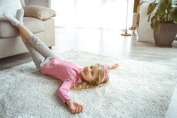 Charming small girl is lying on soft carpet with relaxation in living room. She is looking at...