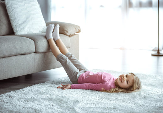 Relaxing at home. Carefree little girl is lying on floor on soft carpet while leaning legs on sofa. She is looking at camera and smiling 