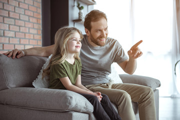 Portrait of kind man is explaining something to his child with gentleness. He is his pointing finger forward and smiling. Girl is sitting on couch next to dad in living room 