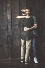 Full length portrait of dark-haired guy with tattoo on hand embracing boyfriend and looking at camera with smile. Young men standing on wooden background