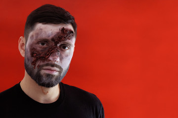 A man with a large torn facial wound on a red background. Portrait of a guy with makeup in the style of Halloween. Copy space.