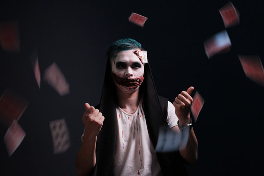 Halloween makeup. The guy in the image of Joker threw cards. Black background. Bloody wounds and drips on the face. Awful illusionist magician.