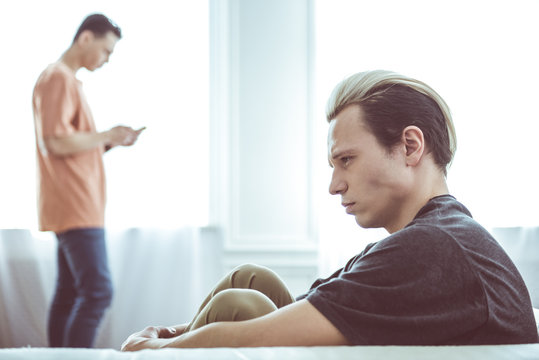 I am ignoring you. Toned side view portrait of sad guy with dyed hair sitting on couch while his boyfriend standing near window and using smartphone on blurred background