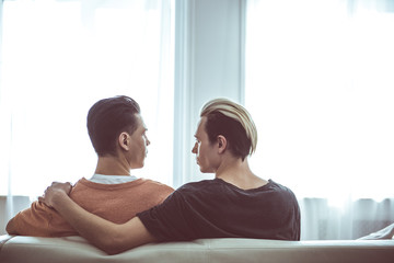 Back view portrait of young man with dyed hair hugging boyfriend and staring at him. They are resting on sofa in front of windows with white curtains