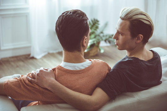 Back view portrait of young man with dyed hair hugging boyfriend and gazing at him. They are resting on sofa at home
