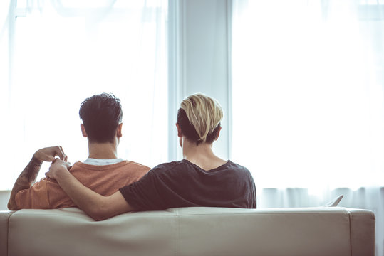 Back view portrait of young man with dyed hair hugging boyfriend while he touching his hand. They are resting on sofa in front of windows