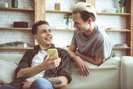 Toned portrait of dark-haired young man sitting on couch and holding cellphone while his boyfriend standing behind. They looking at each other and smiling
