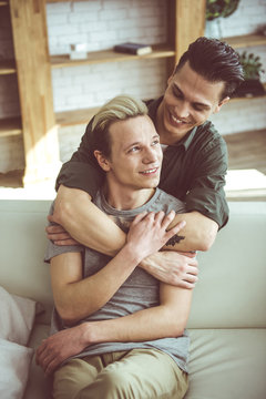 Precious moments with you. Toned vertical portrait of handsome blond guy sitting on couch while his boyfriend hugging him from behind