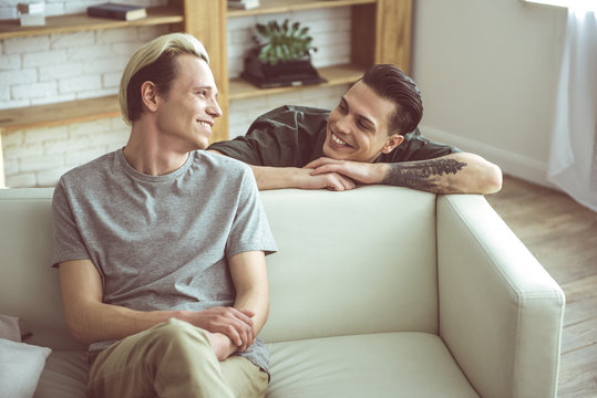 Are you flirting with me. Toned portrait of blond young man sitting on couch while his cheerful boyfriend standing behind and gazing at him