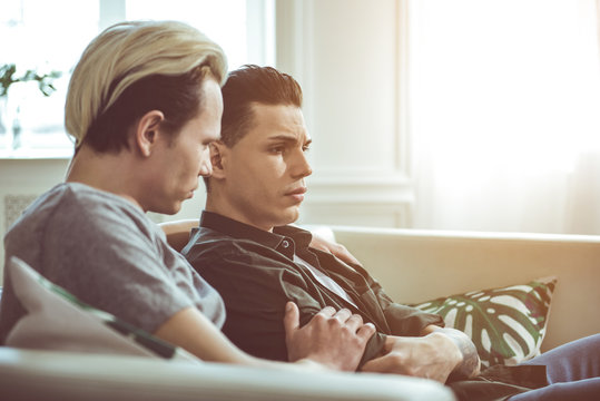 Something wrong. Toned side view portrait of unhappy gay couple sitting on couch at home