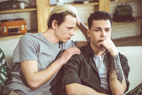 Talk to me. Toned waist up portrait of gay couple sitting on couch at home. Dark-haired guy with tattoo on hand looking away with unhappy expression