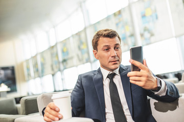 Waist up portrait of man sitting in armchair and holding phone, while having coffee. Copy space on...