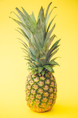 A whole pineapple on a bright yellow background showing the patterned outer skin and healthy leaves.