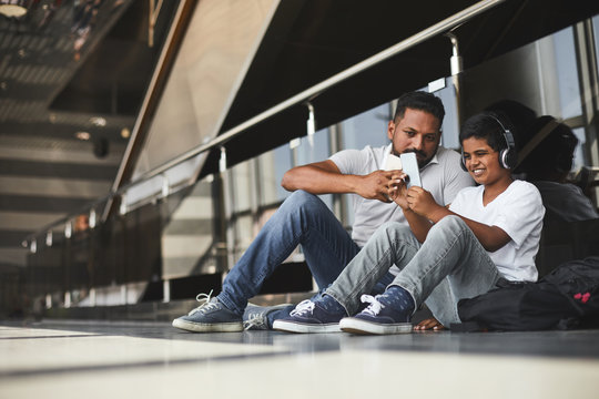 Cheerful little boy sitting on the floor of the airport and playing mobile games while waiting for a flight with his father