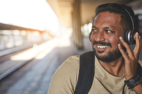 Joyful adult hindu man enjoying music while waiting for a train on the platform