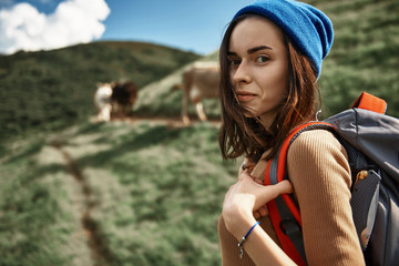 Focus on portrait of grinning woman with backpack standing outdoors. She walking in highland near cow upland pastures