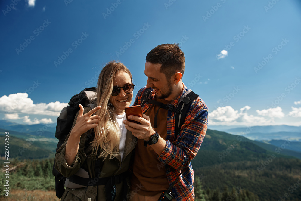 Wall mural Joyful man showing mobile phone to smiling woman while they are standing on top. They having fun together while hiking with backpacks in highland