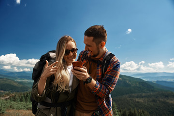 Joyful man showing mobile phone to smiling woman while they are standing on top. They having fun together while hiking with backpacks in highland