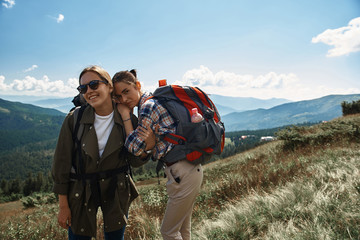 Joyful women hiking together with rucksacks among green tops. They standing and embracing while having great time