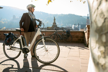 Obraz premium Frau mit Elektro Fahrrad vor Heidelberg Kulisse mit Schloß und alter Brücke , in der Herbst Sonne am Morgen