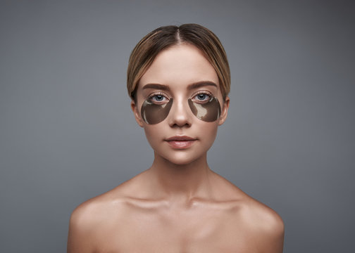 Eye patches. Portrait of the young woman sitting with bare shoulders against the grey background while using eye patches