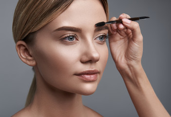 Fototapeta premium Calm young lady standing against the grey background with professional cosmetologist brushing her eyebrows