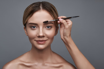 Fototapeta premium Cheerful young lady smiling while sitting against the grey background with the brush near the eyebrow