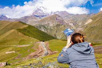 Naklejka premium girl looking in binoscope directed towards the snowy mountain tops of Kazbeg from the view point Gergeti Trinity Church Tsminda Sameba Holy Church near the village of Gergeti in Georgia.