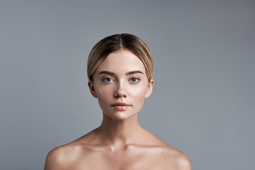 Fototapeta premium Prosty portret. Spokojna śliczna jasnowłosa dama siedząca na szarym tle z nagimi ramionami i lekko uśmiechnięta, patrząc na ciebie