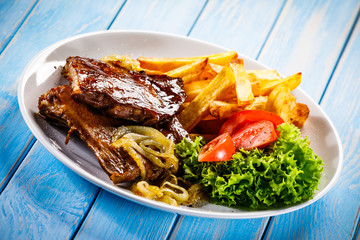 Grilled ribs, French fries and vegetables on wooden background