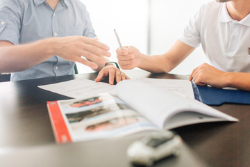 Picture of two men sit at table together. They hold hands on documents. Guy on right gives pen to man on left. There is journal on table. Small white car is in front of picture.
