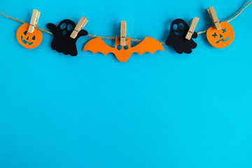 Holiday decorations for Halloween. Black paper ghosts and orange paper pumpkins and bat hanging on a rope on a blue background with copy space.