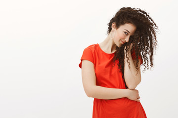 Blushing flirty girlfriend in red dress, covering half of face with curly hair and staring with one eye at camera, smirking, being timid and cute while standing with lowered head over gray wall