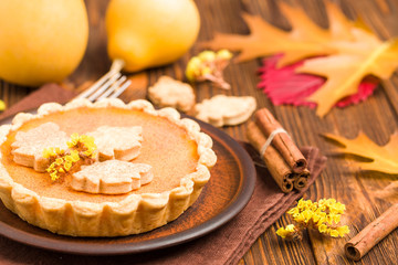 Pumpkin pie with cinnamon and cookies on brown napkins on wooden background with autumn yellow leaves.