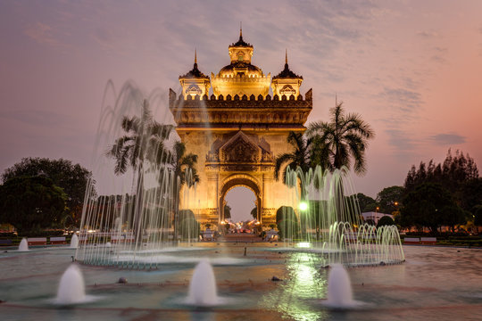Fountain and palm trees in front of the lit Patuxai (Patuxay), Victory Gate or Gate of Triumph, war monument in Vientiane, Laos, at sunset.