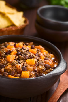 Homemade chili con carne with mincemeat, red and black beans, tomato sauce and pumpkin in rustic bowl with tortilla chips in the back (Selective Focus, Focus in the middle of the dish)
