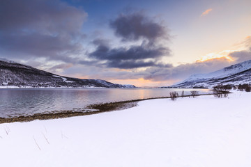 Sunset at the lakeside with rocks of a fjord during low tide in a snowy winter landscape.