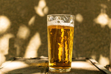 A glass of beer with alcohol. Golden, intoxicating drink in a transparent mug, stands on a wooden stand in the rays of daylight. Top white and airy foam.