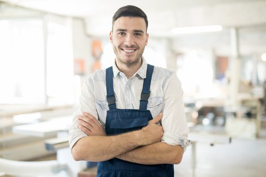 Young successful worker in overalls crossing his arms on chest and looking at you in workshop
