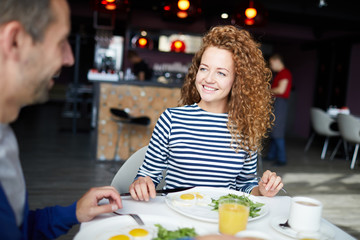 Cheerful young woman with curly hair takling to her colleague or friend by breakfast in cafe