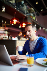 Modern businessman having tea or coffee while sitting by table in front of laptop and networking
