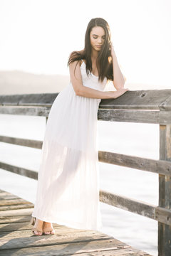 Portrait of a young woman in a white summer dress looking down standing on a pier during the golden hour sunset.