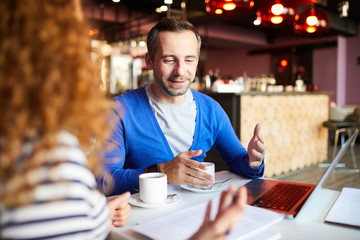 Confident businessman explaining points of project while preparing it with colleague by cup of coffee in cafe
